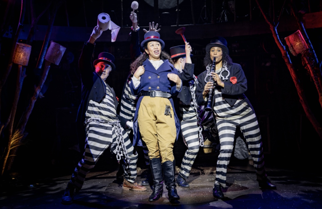 A production shot of Jacoba Williams playing the Carabiniere surrounded by the company in black and white stripes.