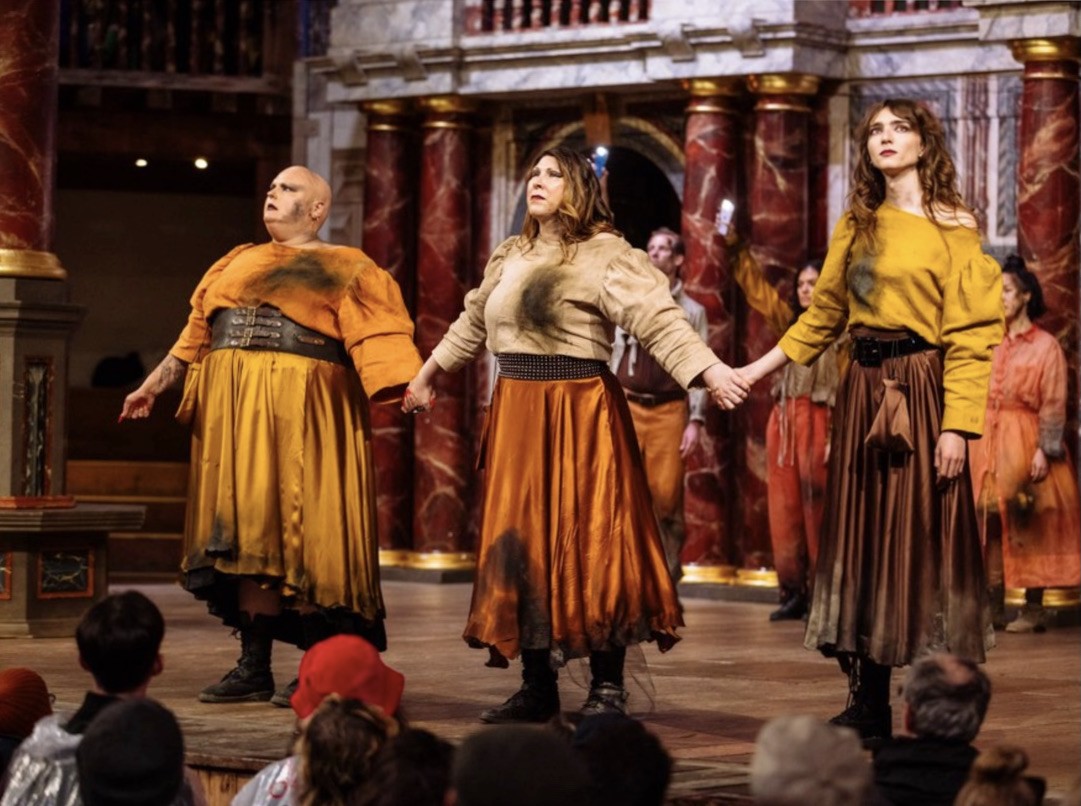A production shot of Burnt at the Stake. 3 actors who played witches in the production in an orange palette of burnt clothing in an orange holding hands.