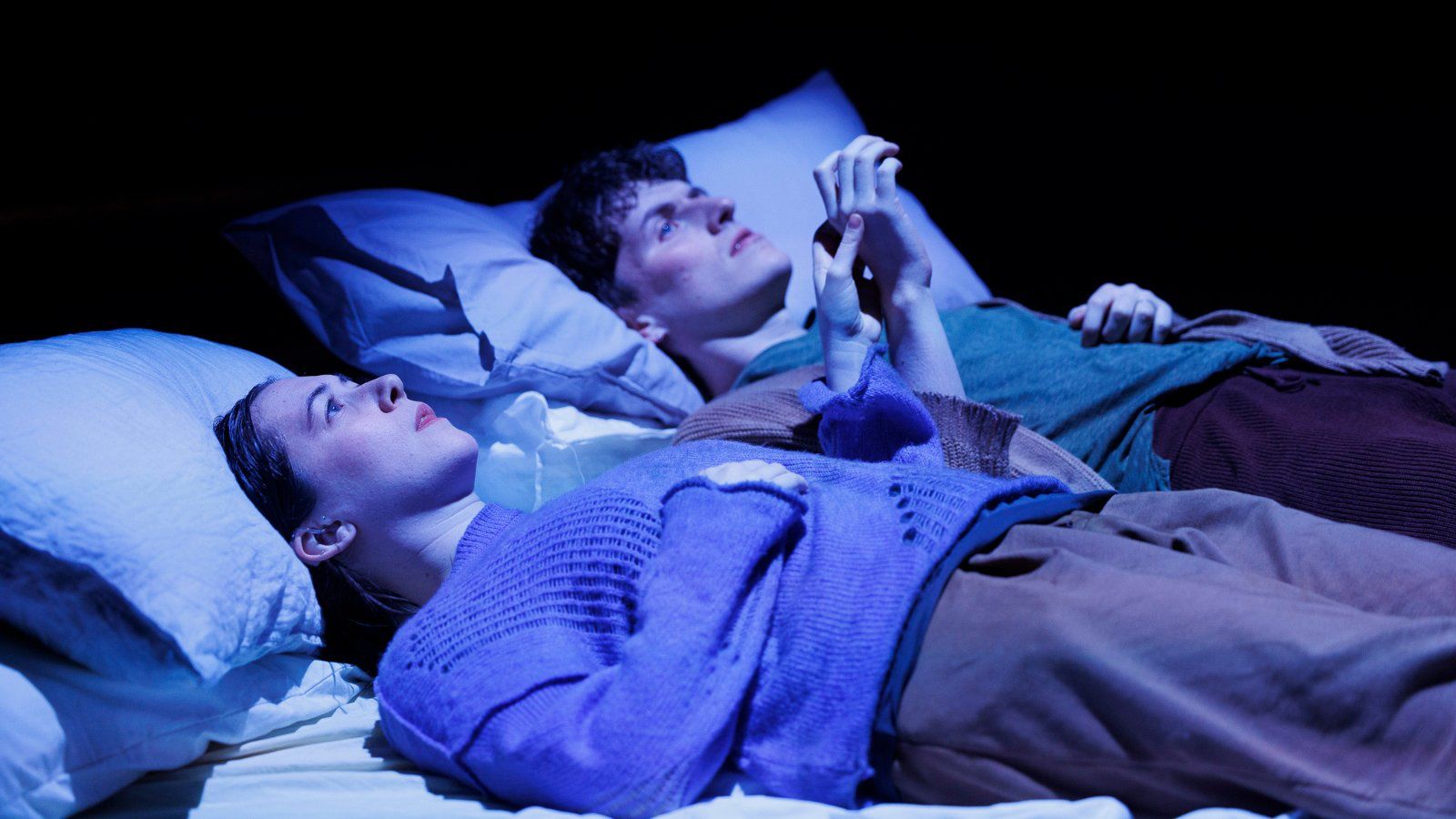 Two people holding hands lying in bed, with a blue light over them.